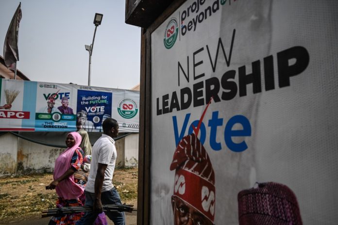 Commuters in Abuja pass a wall covered in election posters. Photo by Dawali David